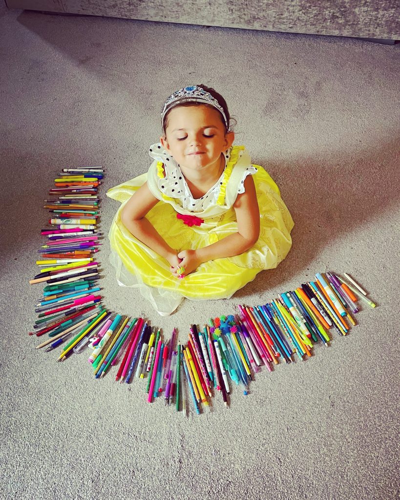 Lilah is pictured from above sat in the middle of a half circle of pens and pencils. She is wearing a yellow princess dress and tiara as she smiles with her eyes closed.