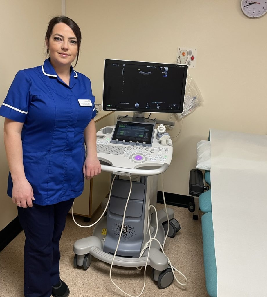 Hayley is stood in a clinical setting to the left of the new ultrasound scanner. The scanner has a large screen at the bottom. Below this is a smaller screen, surrounded by a keyboard. The device has wires around it and is on wheels.