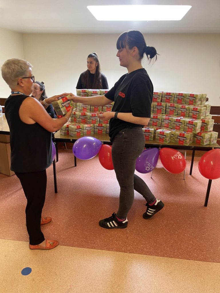 The volunteer is passing a box of tea to a staff member in front of a table filled with hundreds of boxes of tea and red and purple balloons
