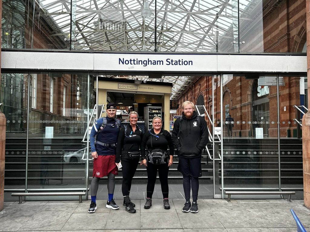 Four people stand in front of the Nottingham Station sign wearing walking clothes and backpacks