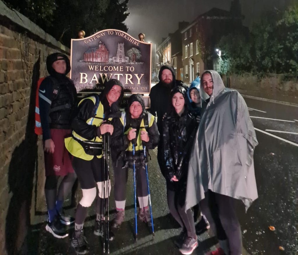 A group of people in walking gear and coats stand in front of the 'Welcome to Bawtry' sign, holding walking sticks. It is dark and raining heavily.